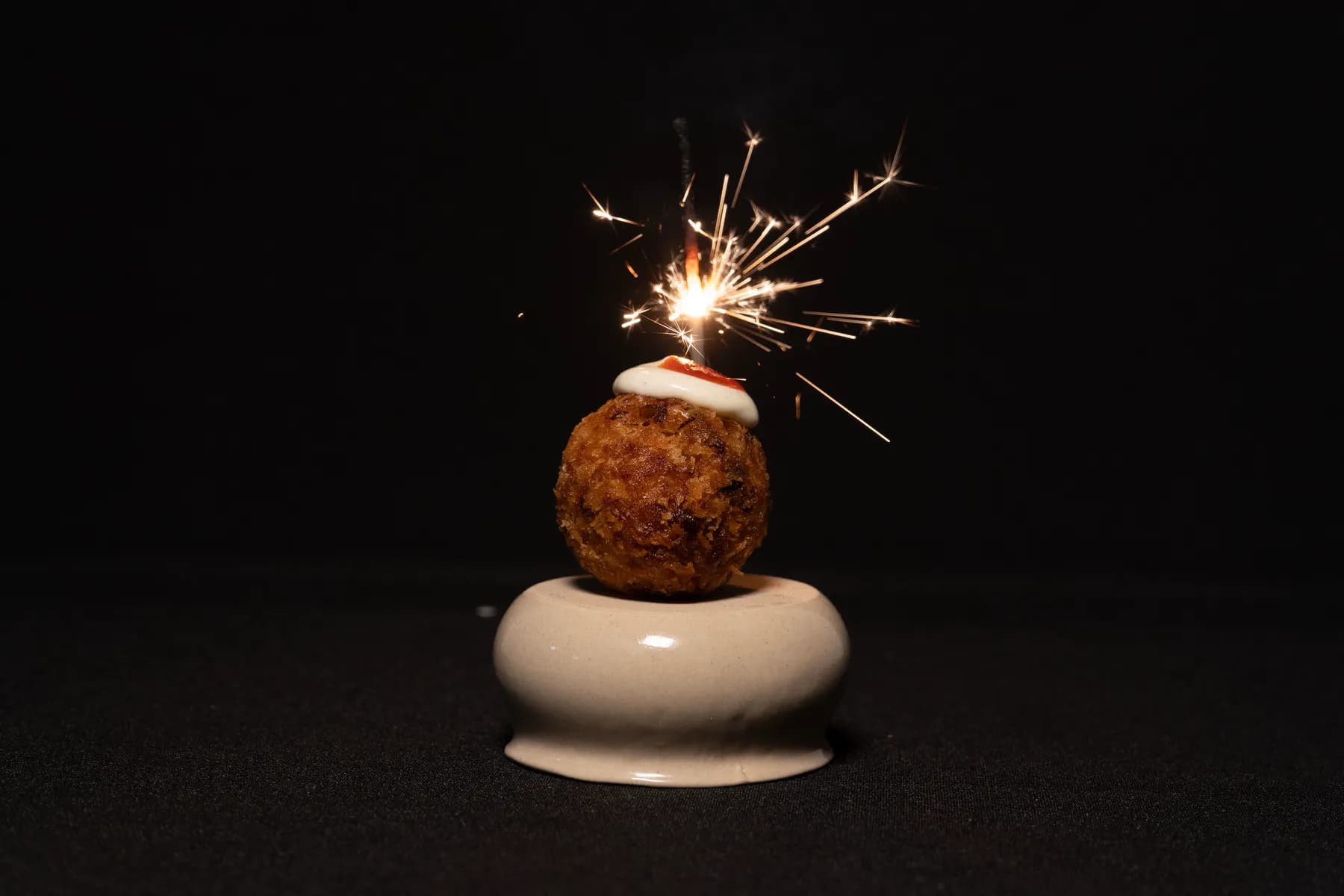 Croqueta artesana de Crocant coronada con alioli y una bengala encendida, sobre fondo negro